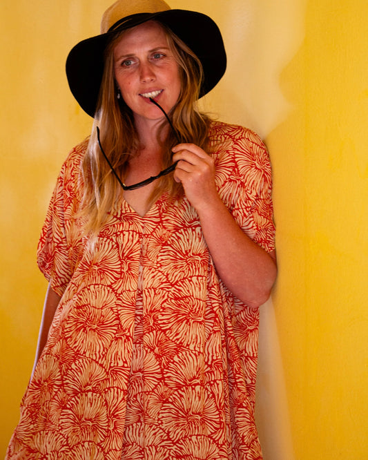 Model wearing the GLORIA Dress in Orange Fizz, posing with a black hat against a yellow background, exuding summer vibes.