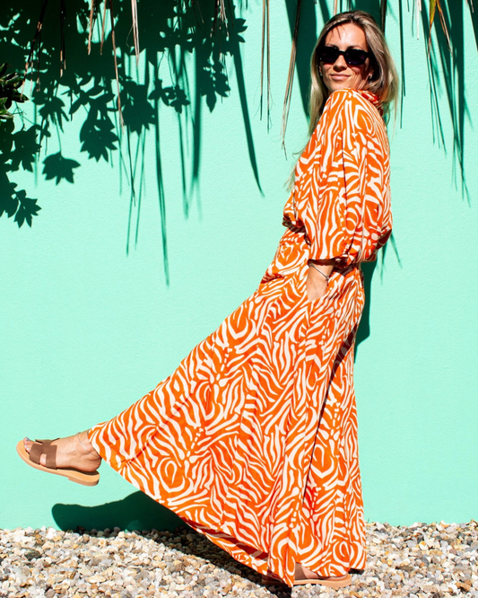 A woman wearing SOFY Pants in a vibrant orange print, standing in front of a turquoise background — The Cocoon Way