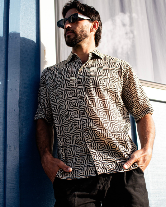 Man wearing SAIRA Short Sleeve Shirt with geometric pattern, sunglasses, and relaxed pose against blue wall.