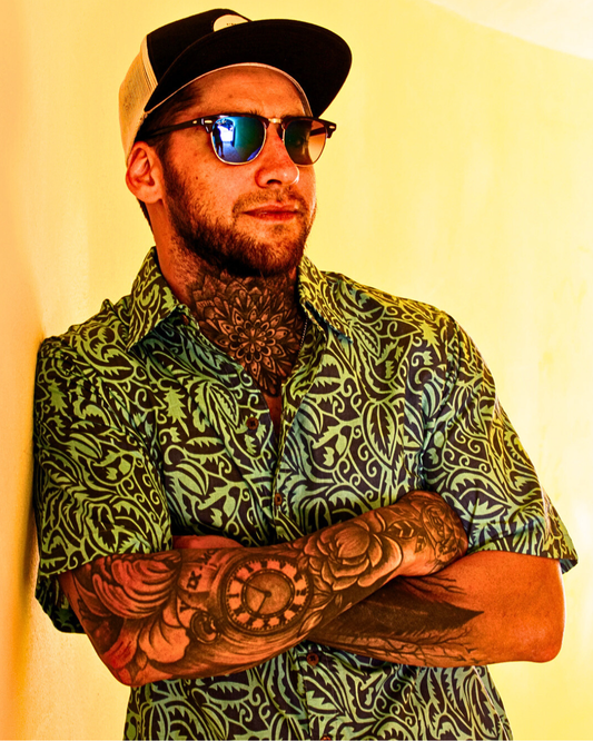 Man wearing a green patterned shirt with tattoos, sunglasses, and a cap, leaning against a wall.