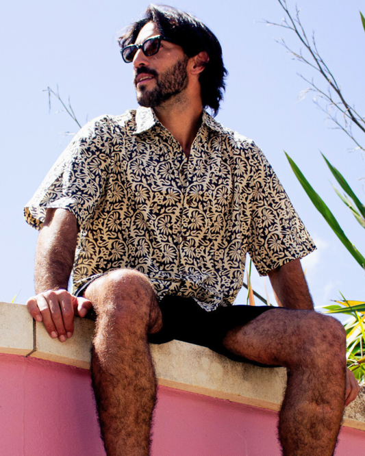 Man wearing SAIRA Short Sleeve Shirt Maui Black sitting on a wall in a sunny outdoor setting — The Cocoon Way