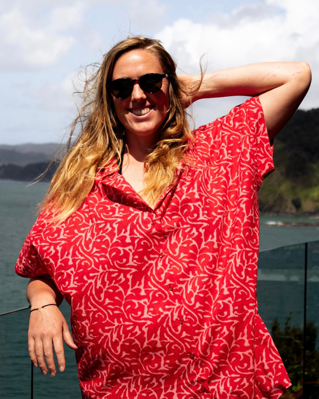 Woman wearing ALBA Maxi Shirt Berry Bloom with sunglasses against scenic coastal background.