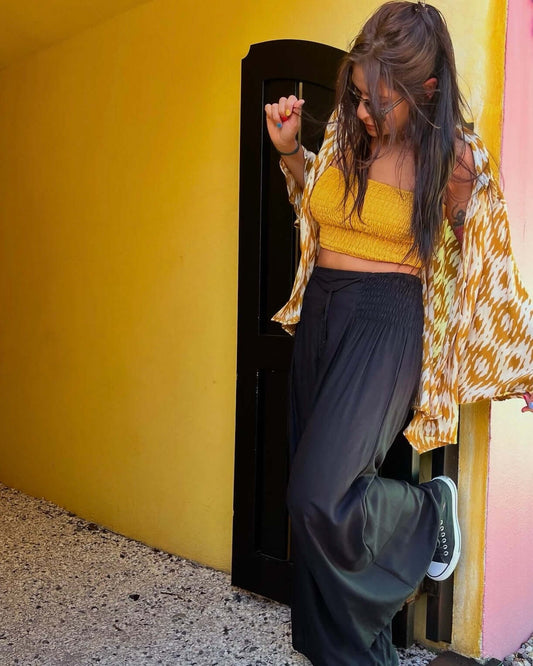 Woman wearing RASA Pants in classic black, styled with a yellow crop top and an open shirt, enjoying a sunny day — The Cocoon Way