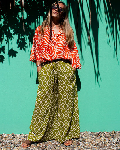 Model wearing OLIVIA Pants in lime ikat pattern with a stylish top and sunglasses against a vibrant green background.
