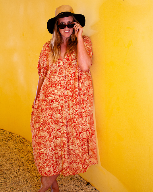 Model wearing GLORIA Dress in Orange Fizz, standing against a bright yellow wall, showcasing a relaxed and stylish look.