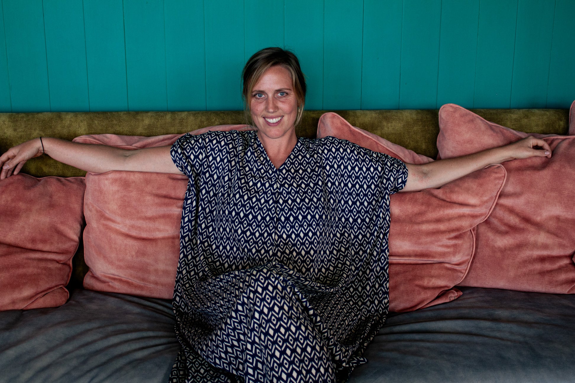 GLORIA Dress worn by a smiling woman sitting on a sofa adorned with pink cushions, showcasing its relaxed design.
