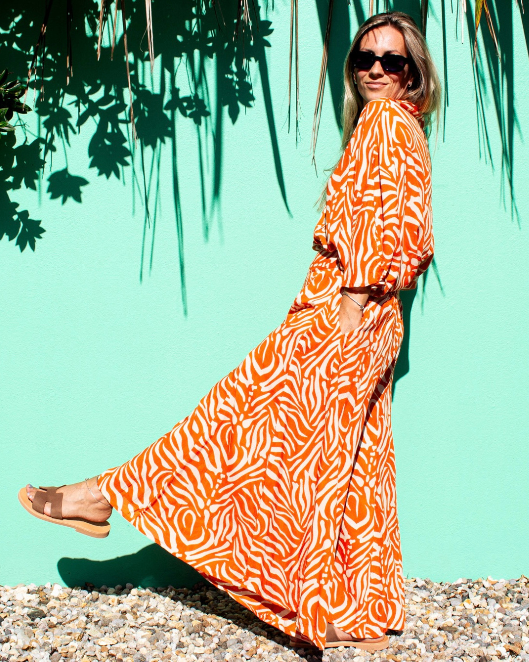 A woman wearing SOFY Pants in a vibrant orange print, standing in front of a turquoise background — The Cocoon Way