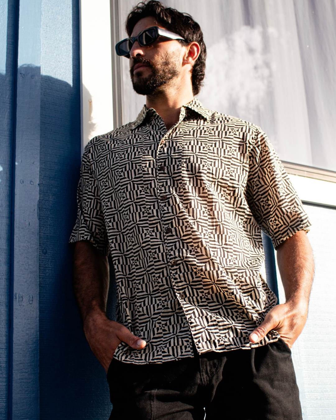 Man wearing SAIRA Short Sleeve Shirt with geometric pattern, sunglasses, and relaxed pose against blue wall.