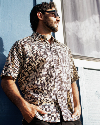 Man wearing SAIRA Short Sleeve Shirt with a bold geometric pattern, standing against a blue wall.