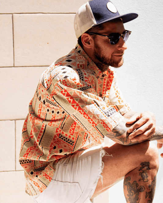 Man wearing the PADMA Short Sleeve Shirt in vibrant orange tribal pattern, paired with sunglasses and a baseball cap — The Cocoon Way