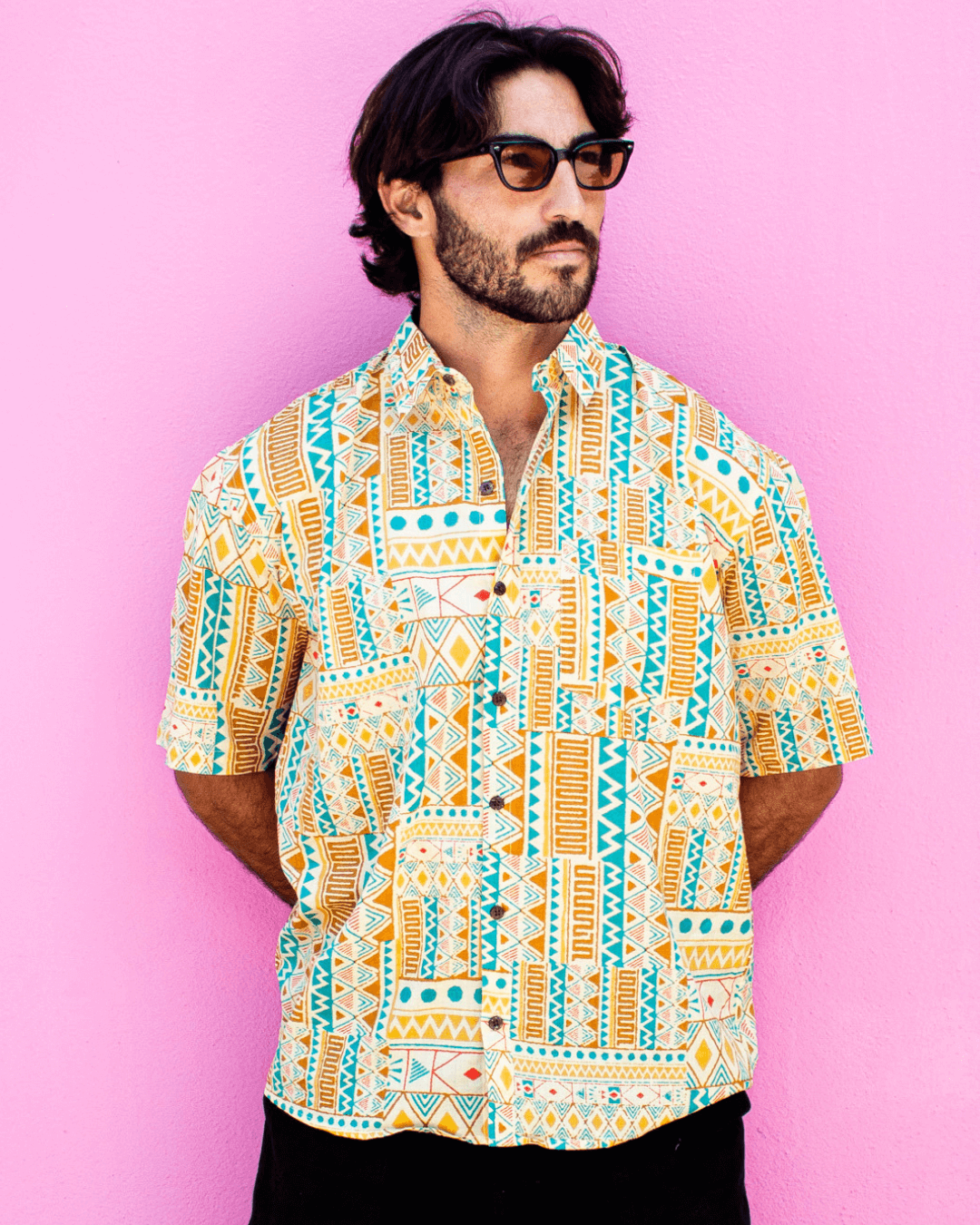 Model wearing PADMA Short Sleeve Shirt in bold yellow tribe print against a pink background.